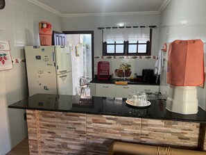 Cozinha com bancada preta, armários brancos, geladeira e janela com cortina de renda.