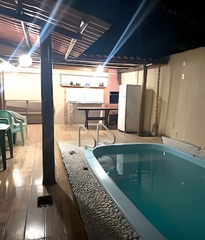An outdoor swimming pool area with a covered seating area and a kitchen in the background. - Residencial Ana e Cesar (Porto Seguro)