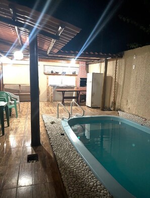 Ein Pool im Freien mit einer gefliesten Liegefläche, einem überdachten Sitzbereich und einer Küche im Hintergrund.