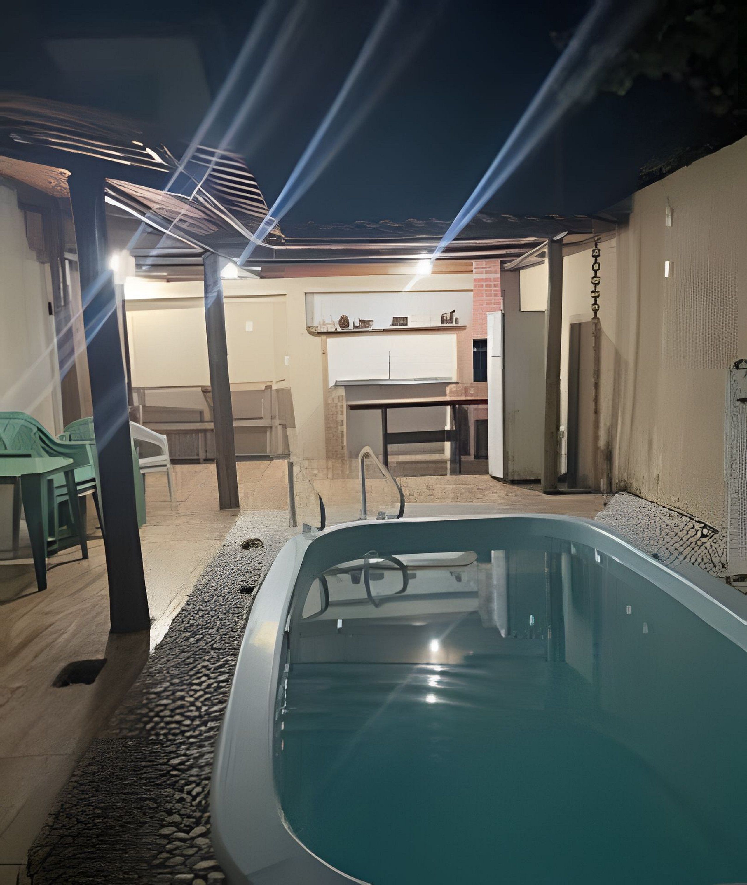A swimming pool with a tiled deck, a covered seating area with chairs, and a kitchen visible in the background.