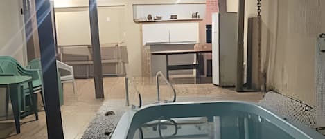 A swimming pool with a tiled deck, a covered seating area with chairs, and a kitchen visible in the background.