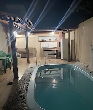 Een zwembad met betegeld zonnedek, een overdekt zitgedeelte met stoelen en een keuken zichtbaar op de achtergrond.