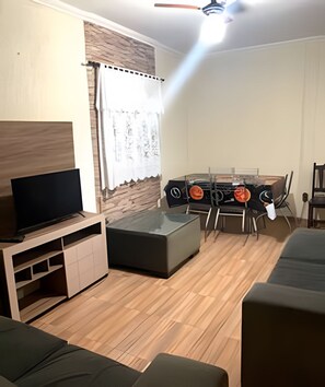 A living room with a flat-screen TV, a sofa, a dining table with chairs, and a ceiling fan.
