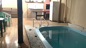 Una piscina con suelo de baldosas, un área cubierta con mesa y sillas, y una cocina al fondo.