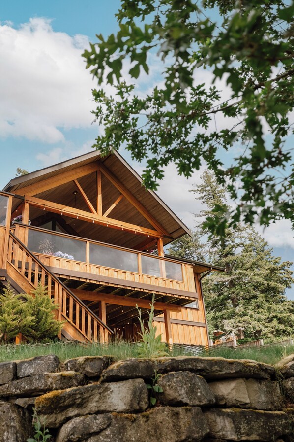 A wooden cabin with a balcony, stone wall, and surrounding trees. - Cabin with Sauna & Cold Plunge, 8 people + kids (Salt Spring Island)