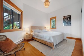 A bedroom with a wooden bed, a nightstand, a chair, a window with blinds, and a poster on the wall. - Cabin with Sauna & Cold Plunge, 8 people + kids (Salt Spring Island)