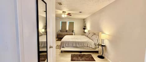 A bedroom with a bed, bedside tables, a ceiling fan, and a view of the adjacent room.