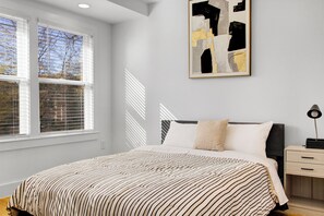 Ein Schlafzimmer mit Bett, Nachttisch, Lampe und einem Fenster mit Jalousien.