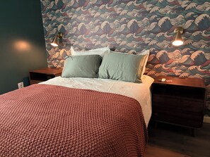 Ein ordentlich gemachtes Bett mit einer roten Strickdecke, zwei Kissen und einer Wand mit Wellenmuster.