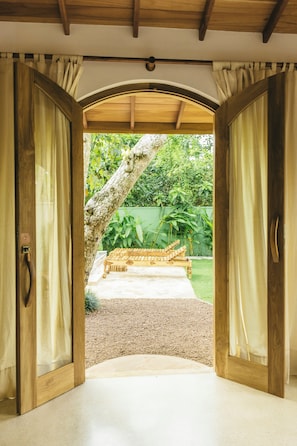 Uma porta de madeira aberta dá para um pátio com um banco de madeira e uma árvore.