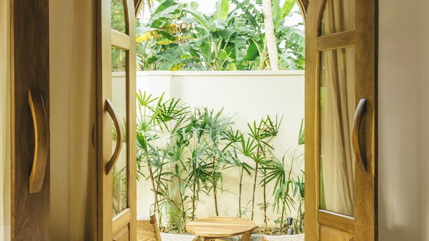 Puerta abierta que da a un patio con una mesa y sillas, rodeado de plantas y vistas al verdor.