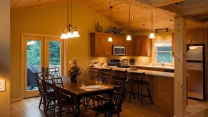 A wooden interior with a dining table, chairs, a kitchen with appliances, and a large window.