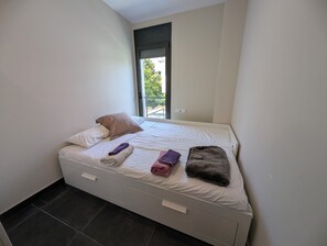 Ein Einzelbett mit Fenster, ein kleiner Nachttisch und ein dunkler Fliesenboden.