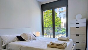 Une chambre moderne avec un grand lit, une table de chevet avec une lampe, une vue sur la verdure par la fenêtre et une commode blanche.