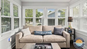 A living room with a beige sofa, a coffee table with remotes, and a window with blinds. - Walk to Lake & Downtown · 3BR Home Sleeps 8 (Lake Geneva)