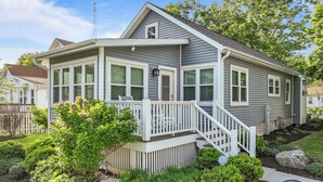 A two-story house with a covered porch, a small staircase, and a landscaped garden with various plants and a large rock. - Walk to Lake & Downtown · 3BR Home Sleeps 8 (Lake Geneva)