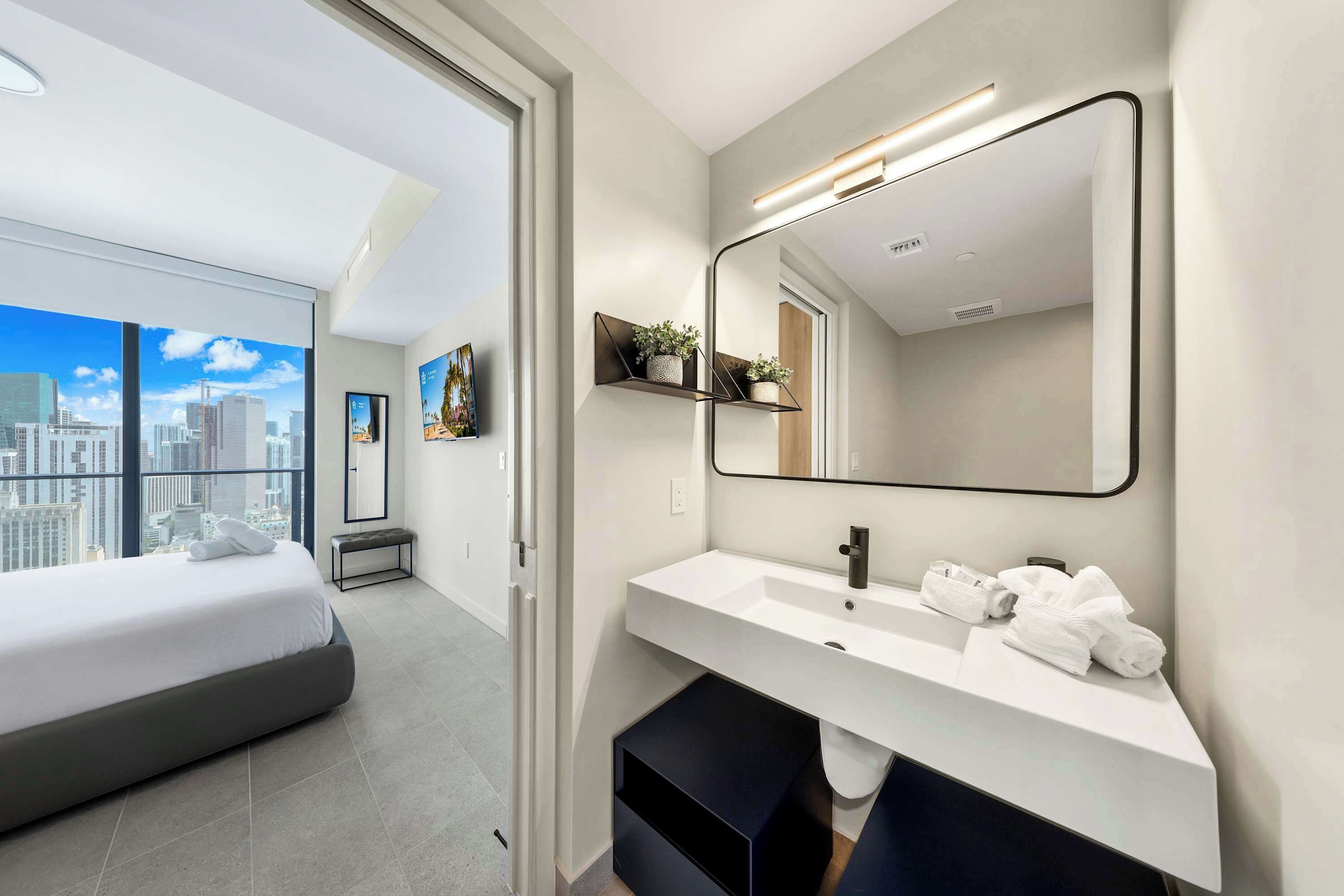 A modern hotel room with a large bed, a bathroom with a white sink and mirror, and a city view.