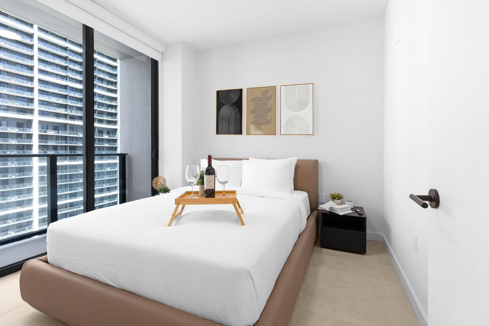 Ein modernes Hotelzimmer mit einem großen Bett, einem Nachttisch, einem Fenster mit Blick auf ein Hochhaus und einem kleinen Beistelltisch.