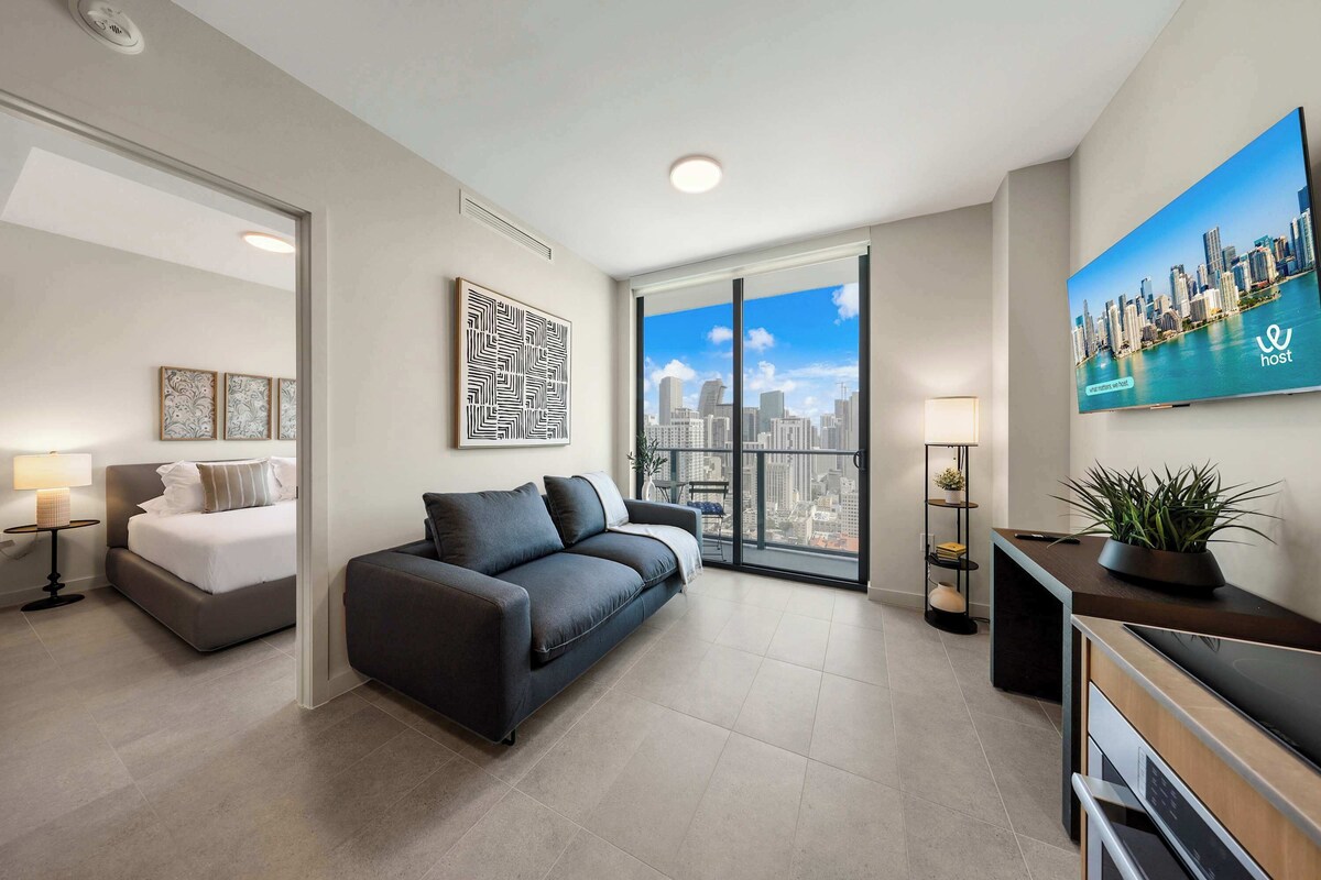 Una sala moderna con cama, sofá, televisor y un amplio ventanal con vistas a la ciudad.