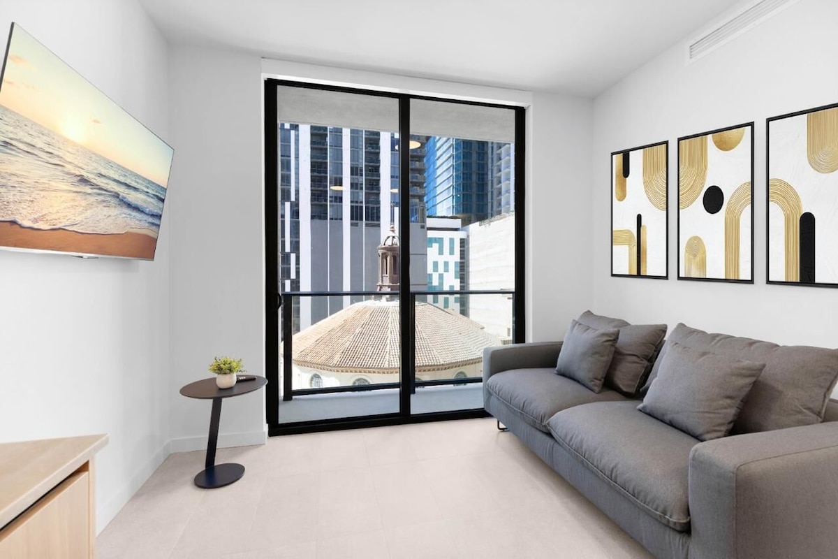 A modern living room with a grey sofa, a small round table with a plant, and a large window with a city view.