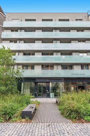 Ein mehrstöckiges Gebäude mit moderner Architektur, das einen zentralen Eingang mit Glastüren, einen gepflasterten Weg und eine Bank bietet.