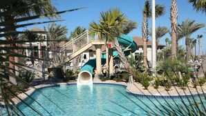 A pool with a water slide, palm trees, and a building with a balcony. - 5BR ChampionsGate Home with Pool & Water Park (Championsgate)