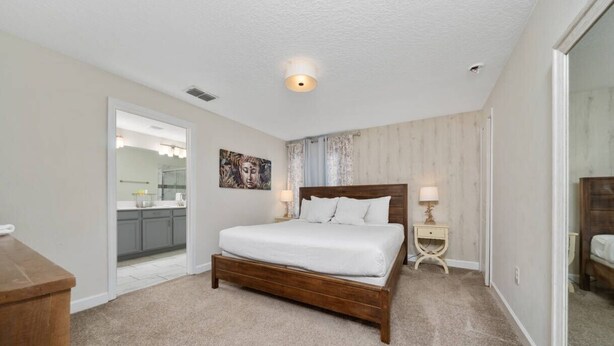 A bedroom with a wooden bed, a nightstand, a bedside lamp, a large mirror, and a view into the kitchen. - 5BR ChampionsGate Home with Pool & Water Park (Championsgate)