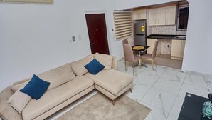Olohuoneessa on beige sohva, ruokapöytä tuoleineen sekä keittiö, jossa on kaappeja ja ikkuna.