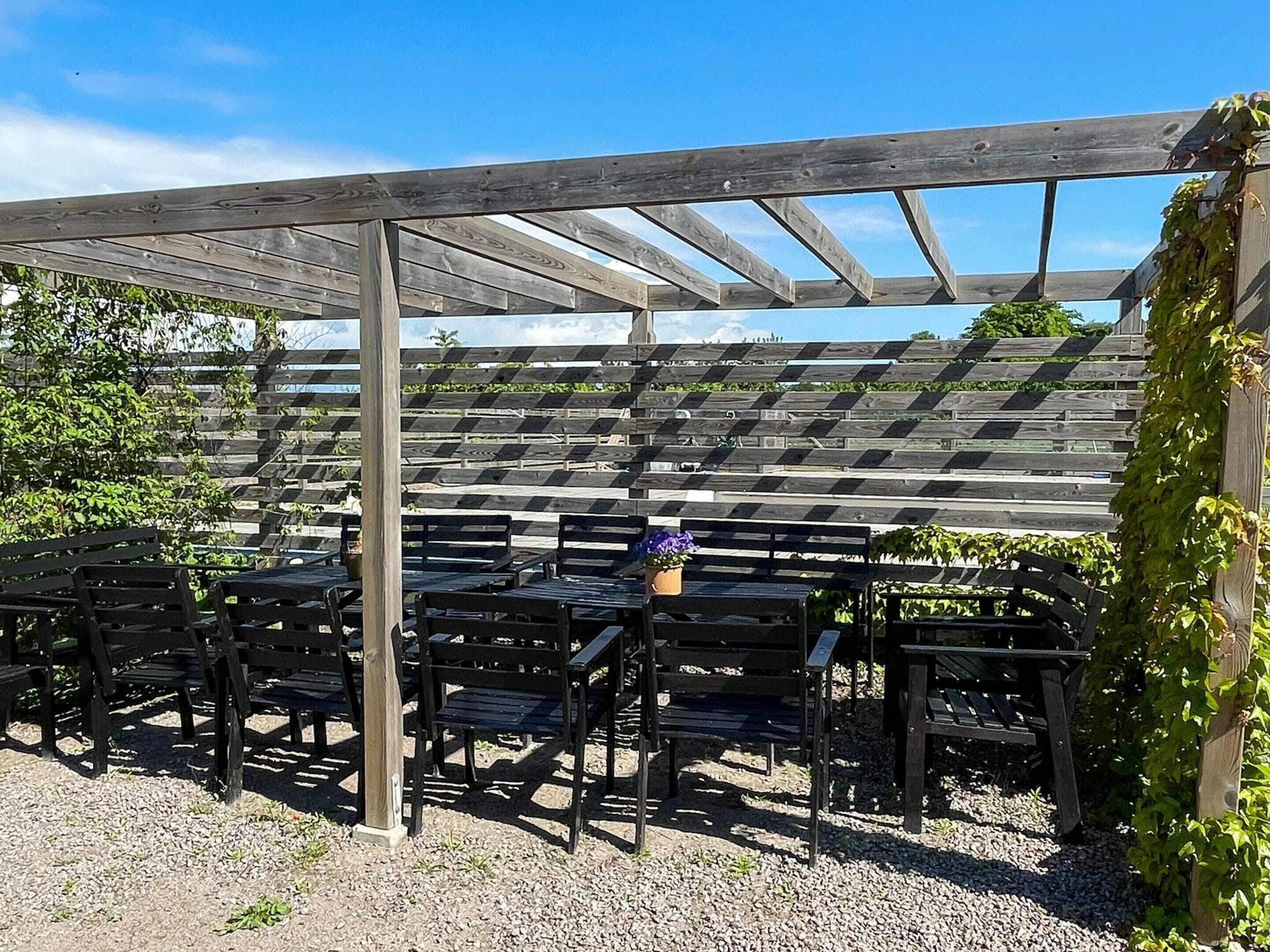 Un área de estar al aire libre con techo, con bancos y mesas de madera, rodeada por una estructura de pérgola de madera.