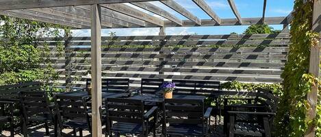 Un área de estar al aire libre con techo, con bancos y mesas de madera, rodeada por una estructura de pérgola de madera.