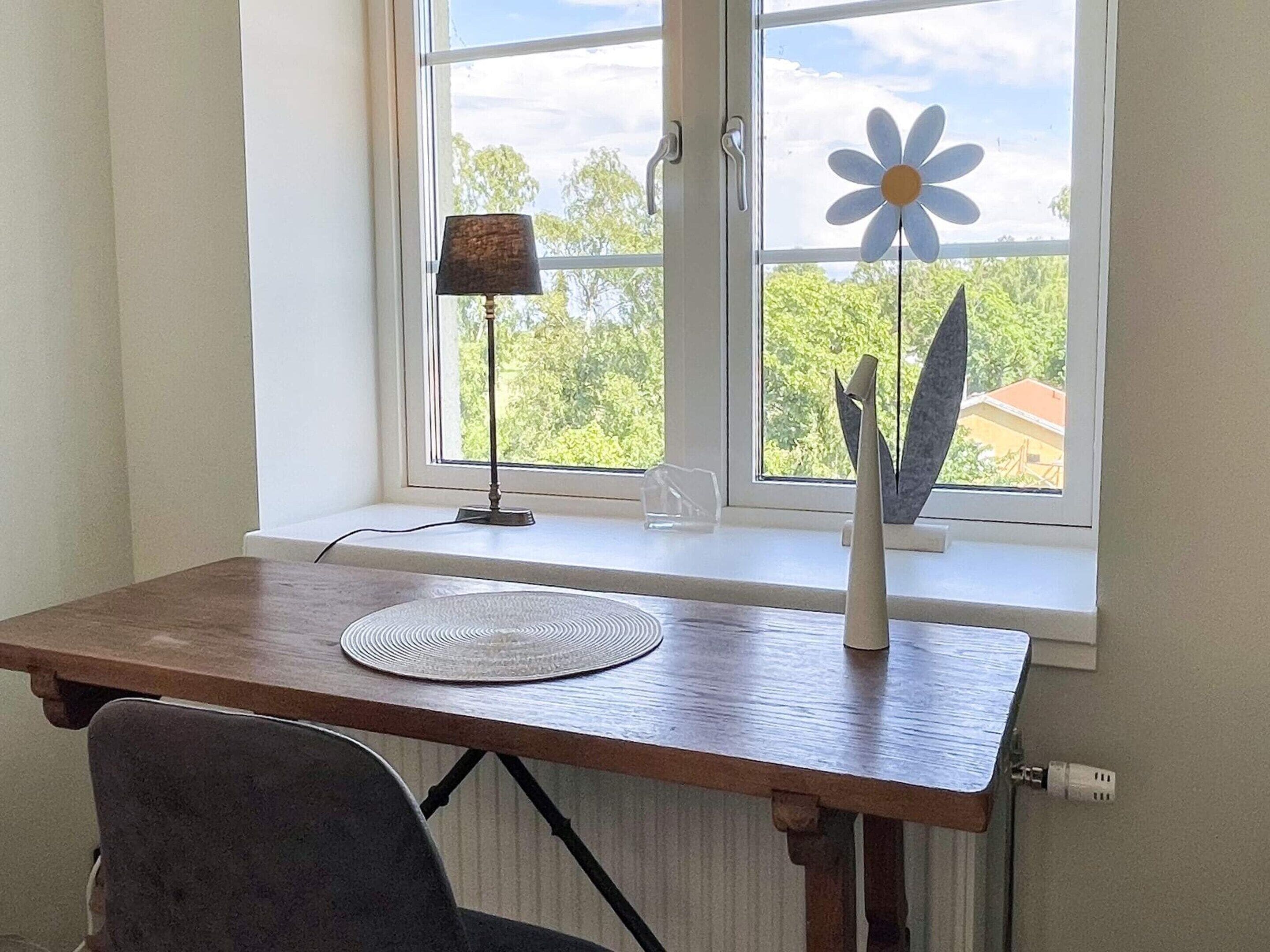 A wooden desk with a lamp, a decorative flower, and a clear view of greenery outside.