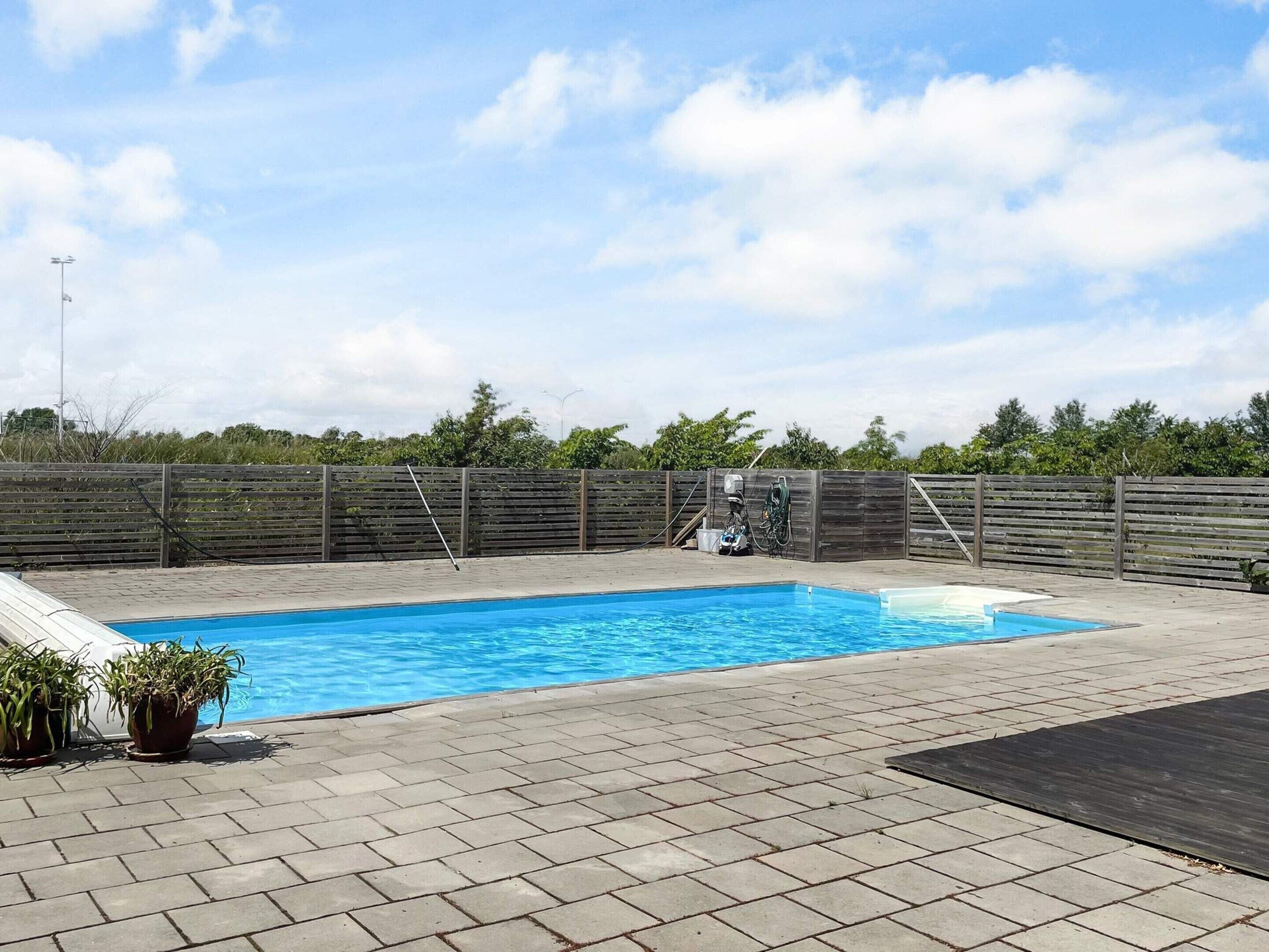 A rectangular swimming pool with a wooden deck surrounding it, a wooden fence, and potted plants.
