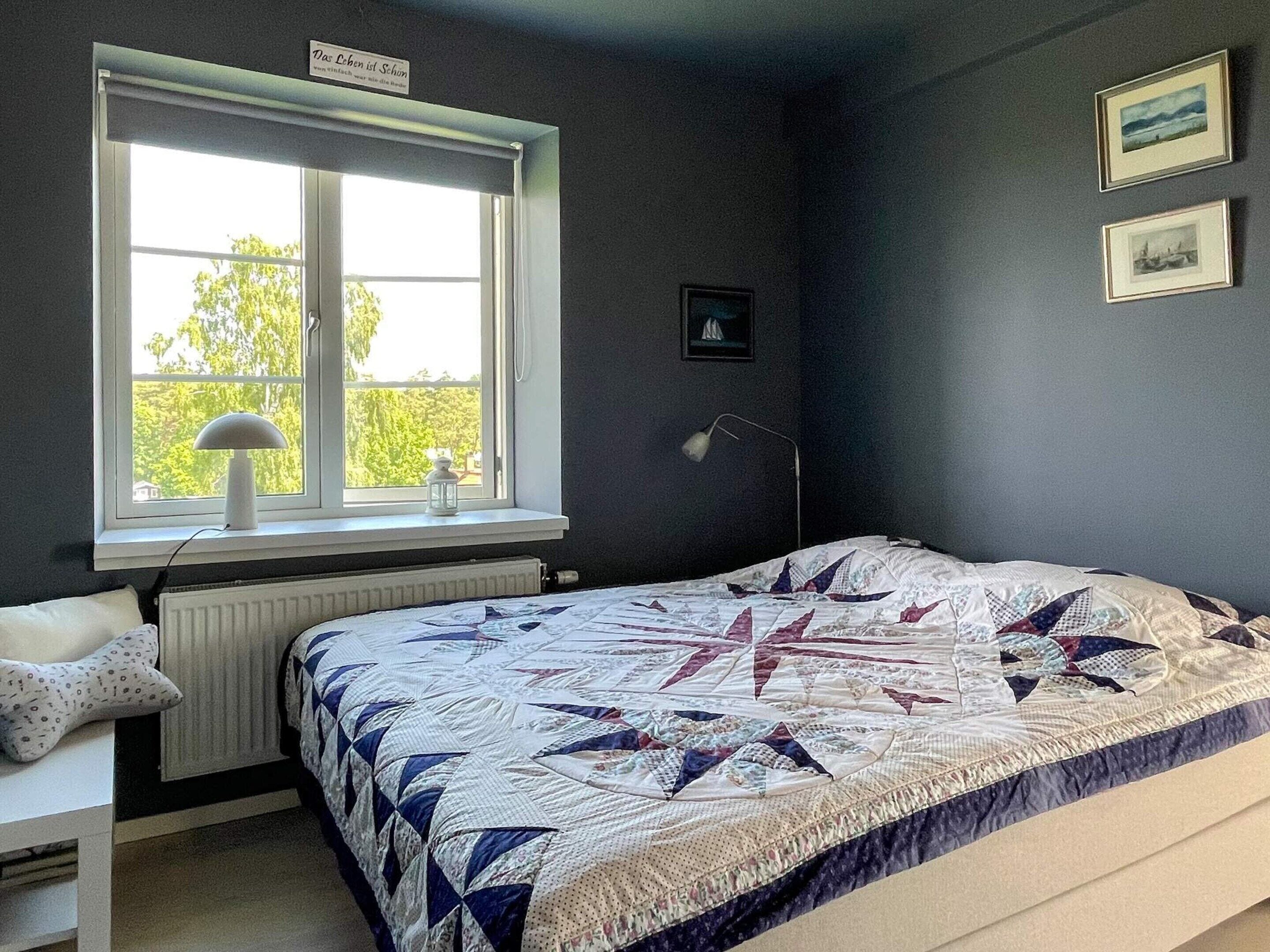 A bedroom with a large window, a radiator, a bed with a quilt, a desk with a lamp, and two framed pictures on the wall.