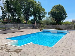 Ein rechteckiger Swimmingpool mit einer gefliesten Liegefläche, umgeben von einem Holzzaun und Bäumen.