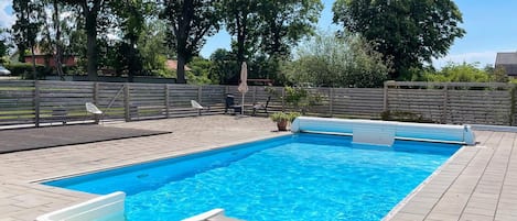 Ein rechteckiger Swimmingpool mit einer gefliesten Terrasse, umgeben von einem Holenzaun und Bäumen.