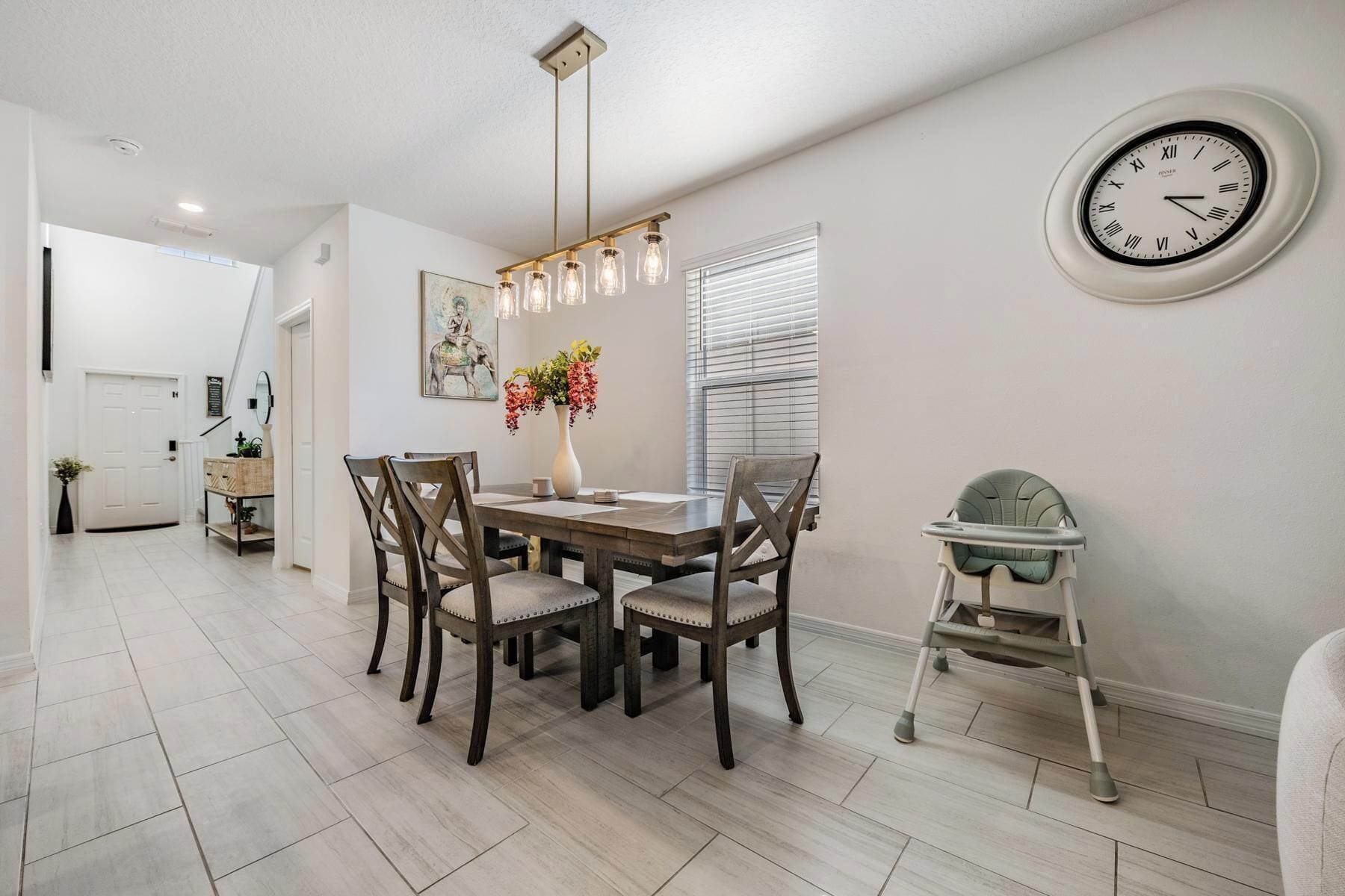 A dining area with a wooden table, four chairs, a vase with flowers, a wall clock, and a baby walker.
