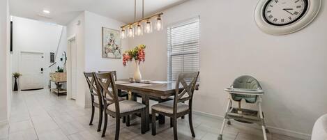 A dining area with a wooden table, four chairs, a vase with flowers, a wall clock, and a baby walker.
