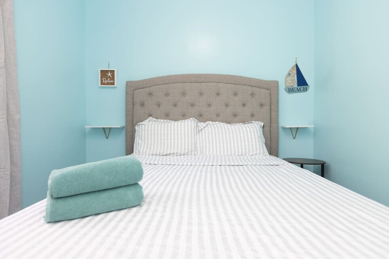 A bed with striped bedding, two pillows, and a folded towel. There are two bedside tables, one with a small decorative item and another with a sign.