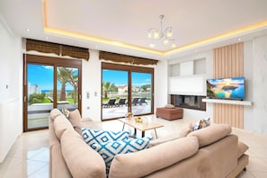 Ein modernes Wohnzimmer mit einer großen Couch, einem Couchtisch und einem an der Wand montierten Fernseher. Es gibt Glastüren, die auf einen Balkon mit Gartenmöbeln führen.