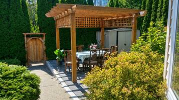 Una pérgola de madera con una mesa y sillas debajo, rodeada de abundante vegetación y un cielo despejado.