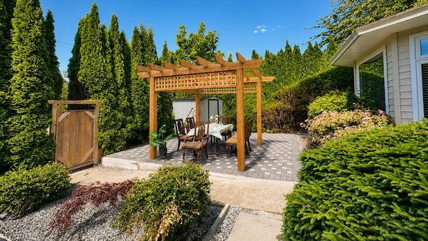 A wooden pergola with a table and chairs, surrounded by well-maintained hedges and trees.