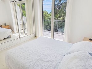 Ein Schlafzimmer mit einem großen Bett, einem Nachttisch, einem Fenster mit Blick auf Bäume und einem Balkon.