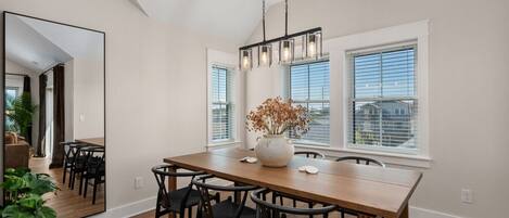 Un comedor con una mesa de madera, sillas negras, un jarrón con flores secas y ventanas amplias.
