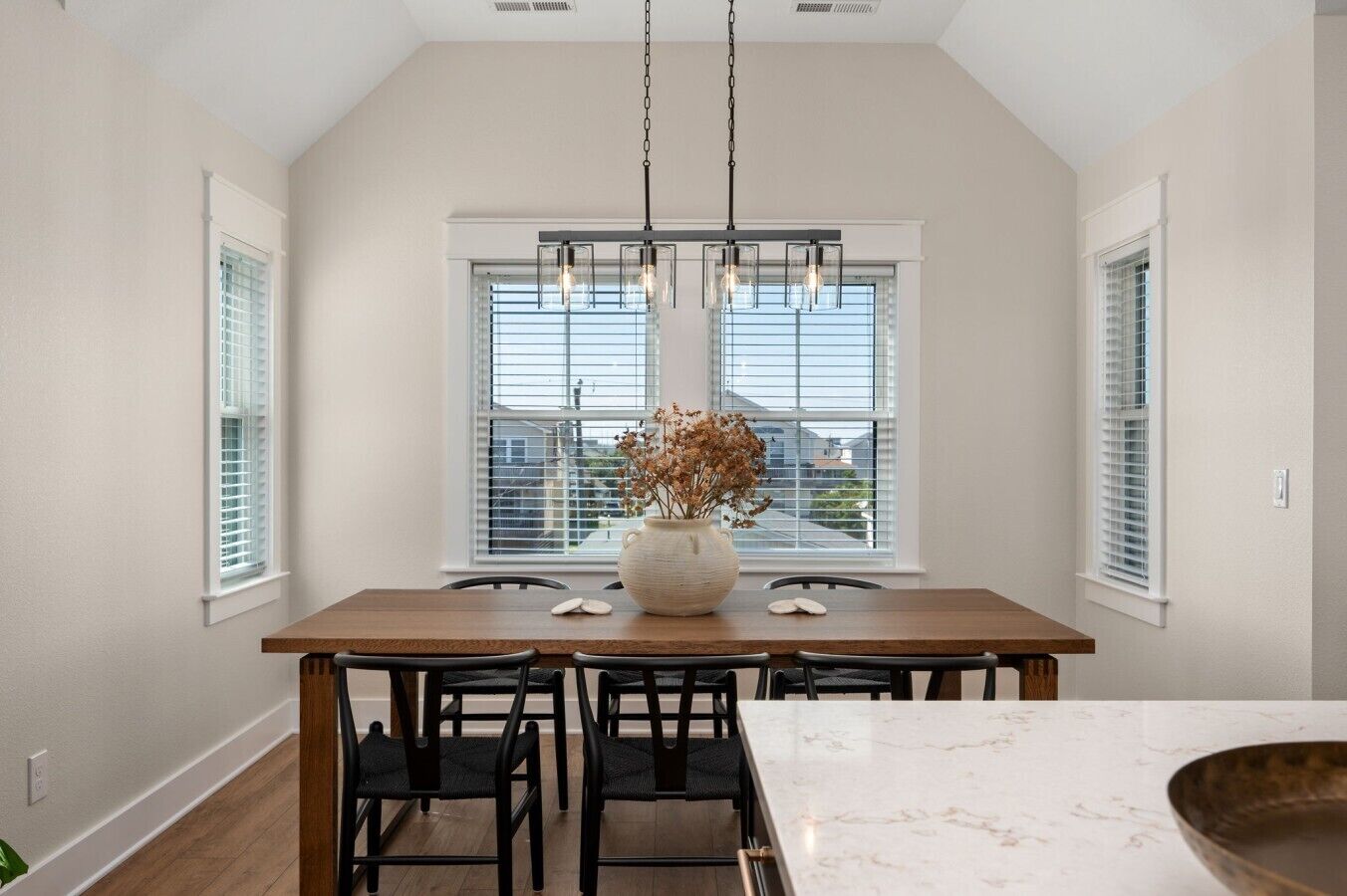 A dining area with a wooden table, chairs, a vase with dried flowers, and a hanging light fixture.
