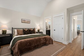 A bedroom with a bed, nightstands, a lamp, a painting, and a door leading to another room. - Luxury Home! Steps To Beach - Pool - Dog Friendly! (Kill Devil Hills)