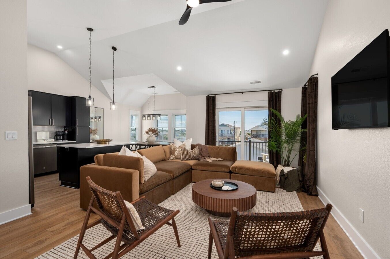 A modern living room with a sectional sofa, a dining table, wicker chairs, a kitchen area, and a large window offering a view.