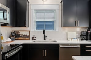 A modern kitchen with dark cabinets, a stainless steel range hood, a built-in dishwasher, and a coffee maker. - Luxury Home! Steps To Beach - Pool - Dog Friendly! (Kill Devil Hills)