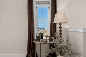 Ein Eckzimmer mit Fenster, einer Lampe, einem kleinen Tisch mit Büchern und einer Kerze sowie einer Topfpflanze.