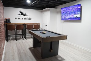 A pool table with chairs, a wall-mounted TV, and a sign reading 'Welcome to the Hangar Hangout.'