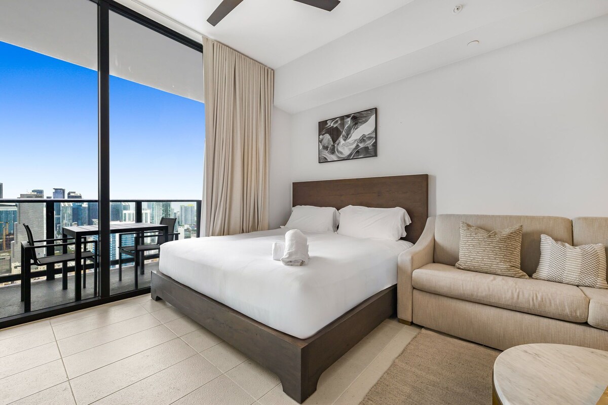 Habitación de hotel moderna con una cama grande, un sofá, una mesa de comedor y vistas a la ciudad.